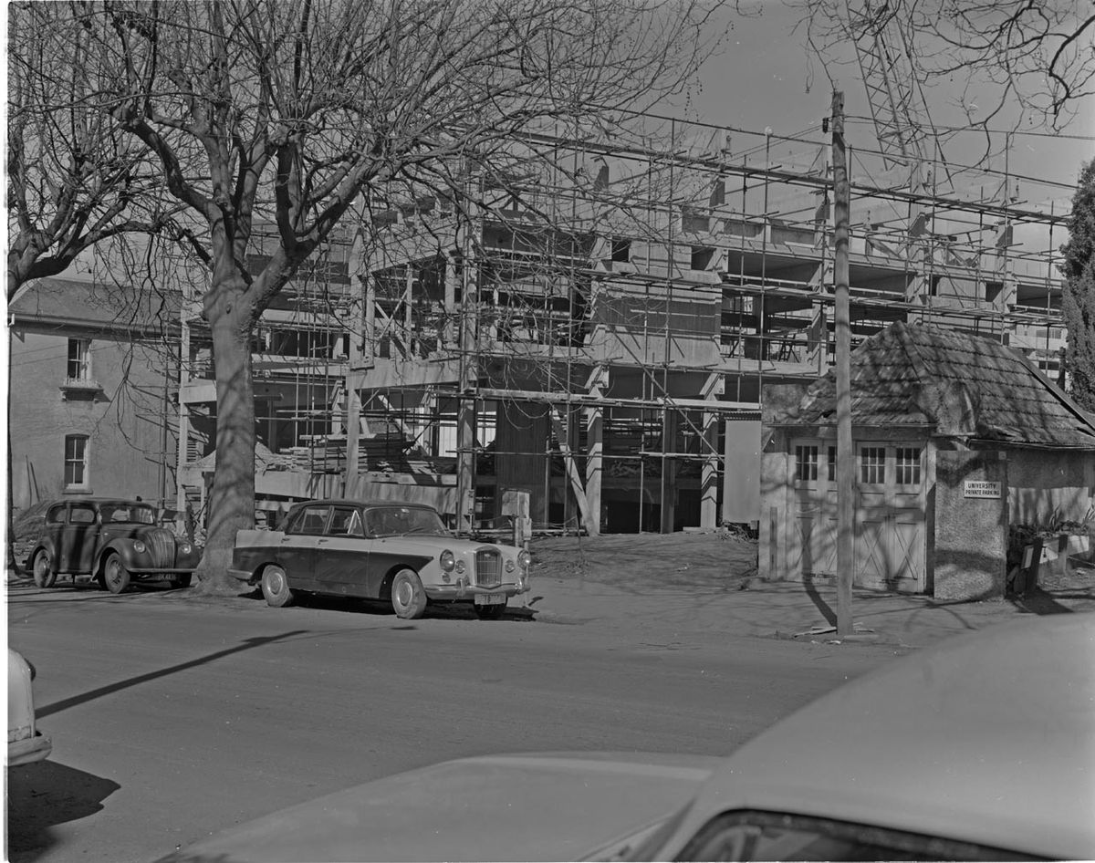 Black-and-white photo of the Student Union Building under construction in the late 1960s, wrapped in scaffolding behind bare trees and parked vintage cars