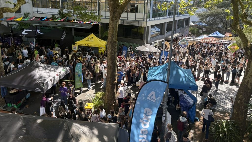 Crowds of students at stalls and tents in the Quad during AUSA's O-Week festival, with an AUSA flag in the foreground